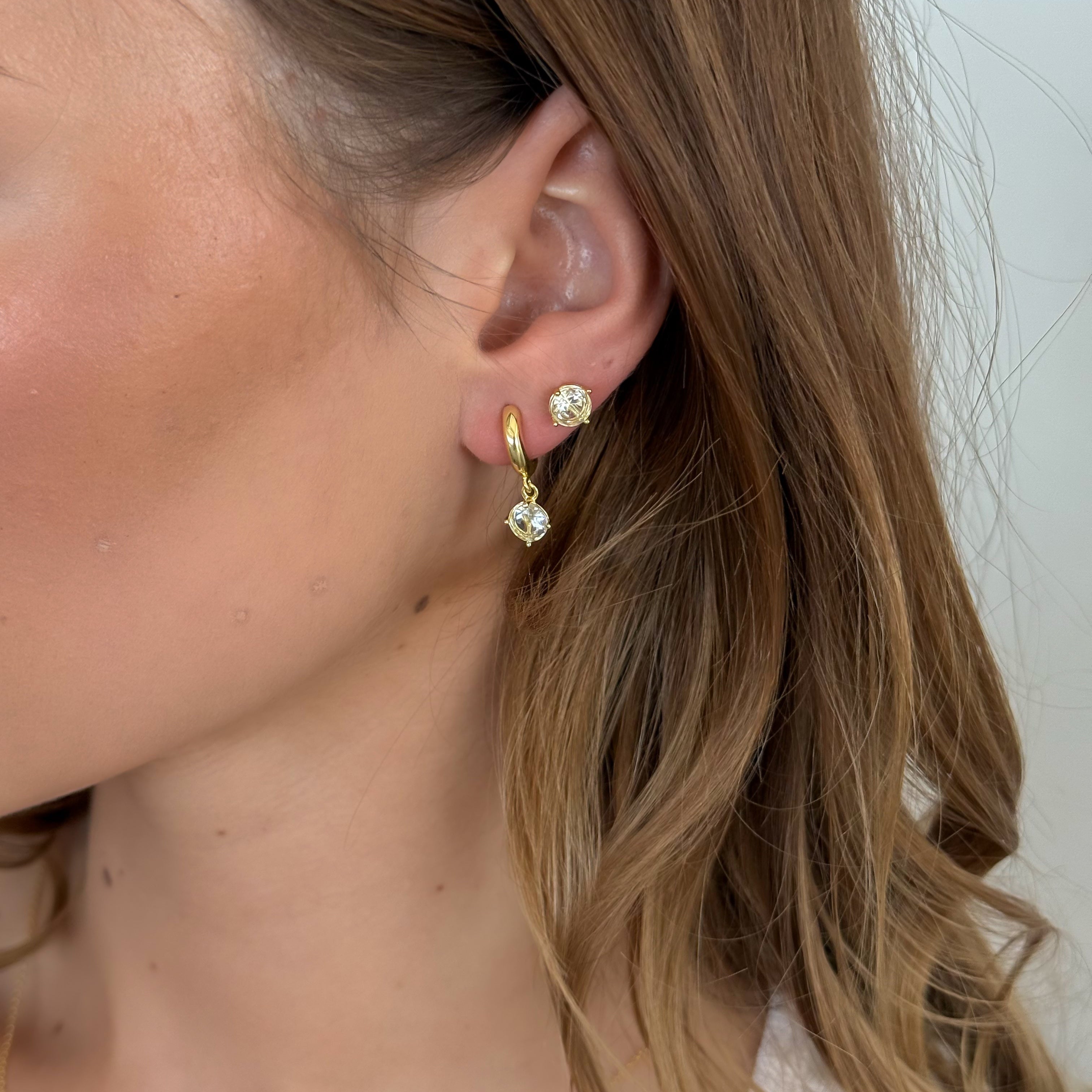 Close-up of a person wearing gold earrings with clear gemstones against a neutral background